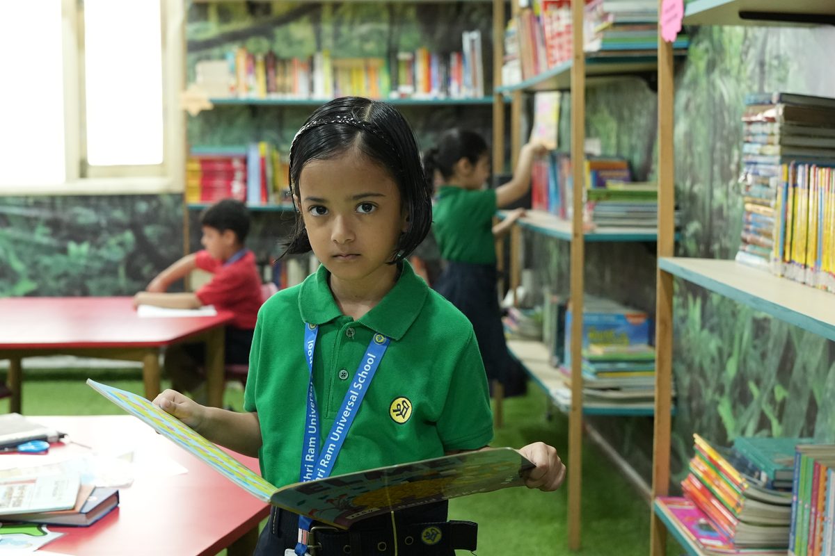 A young girl in a green school T-shirt holding an open picture book in the library at The Shri Ram Universal School, Sarabha Nagar
