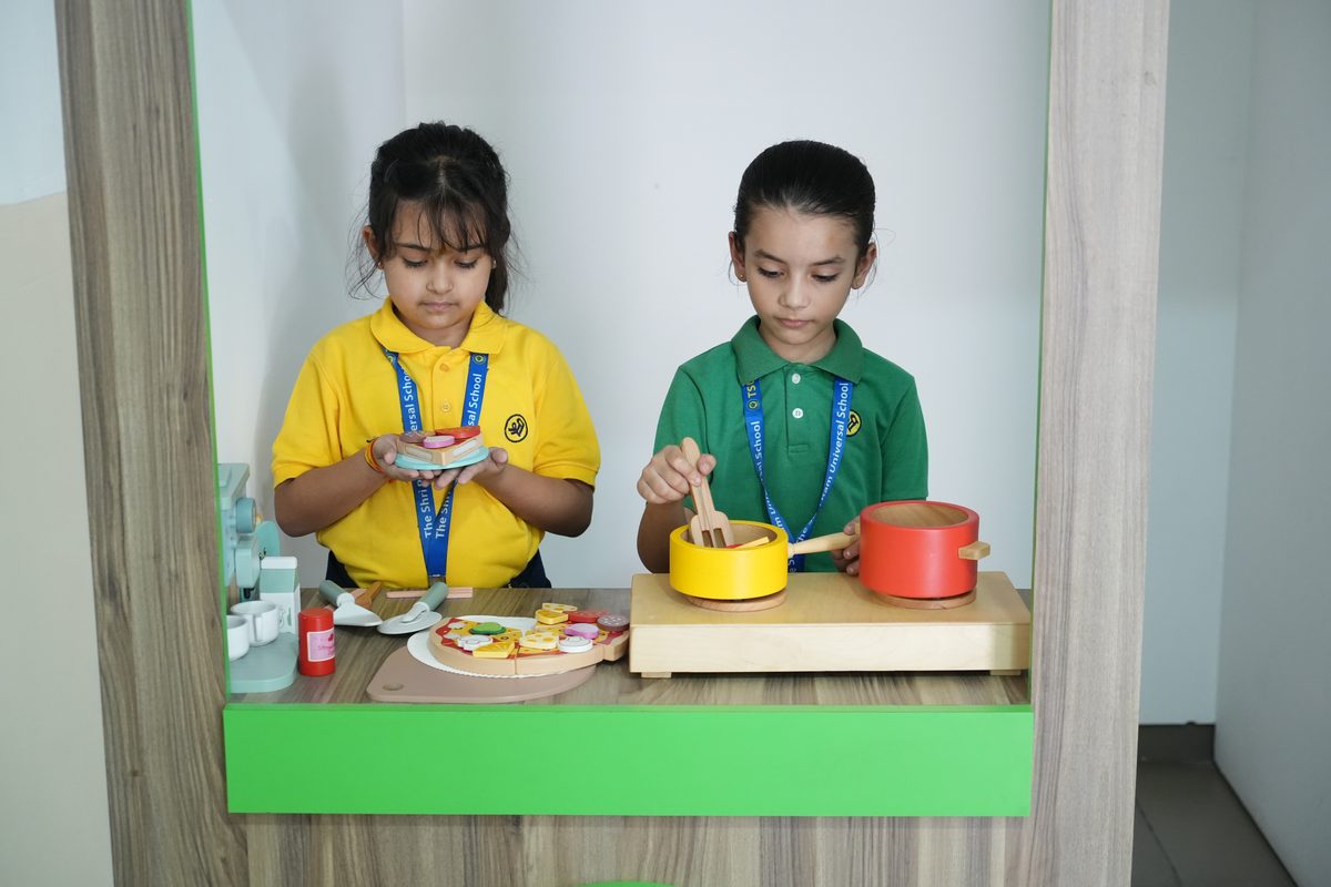 Two young girls cooking together with wooden pots and a toy pizza in the role-play kitchen at The Shri Ram Universal School, Sarabha Nagar