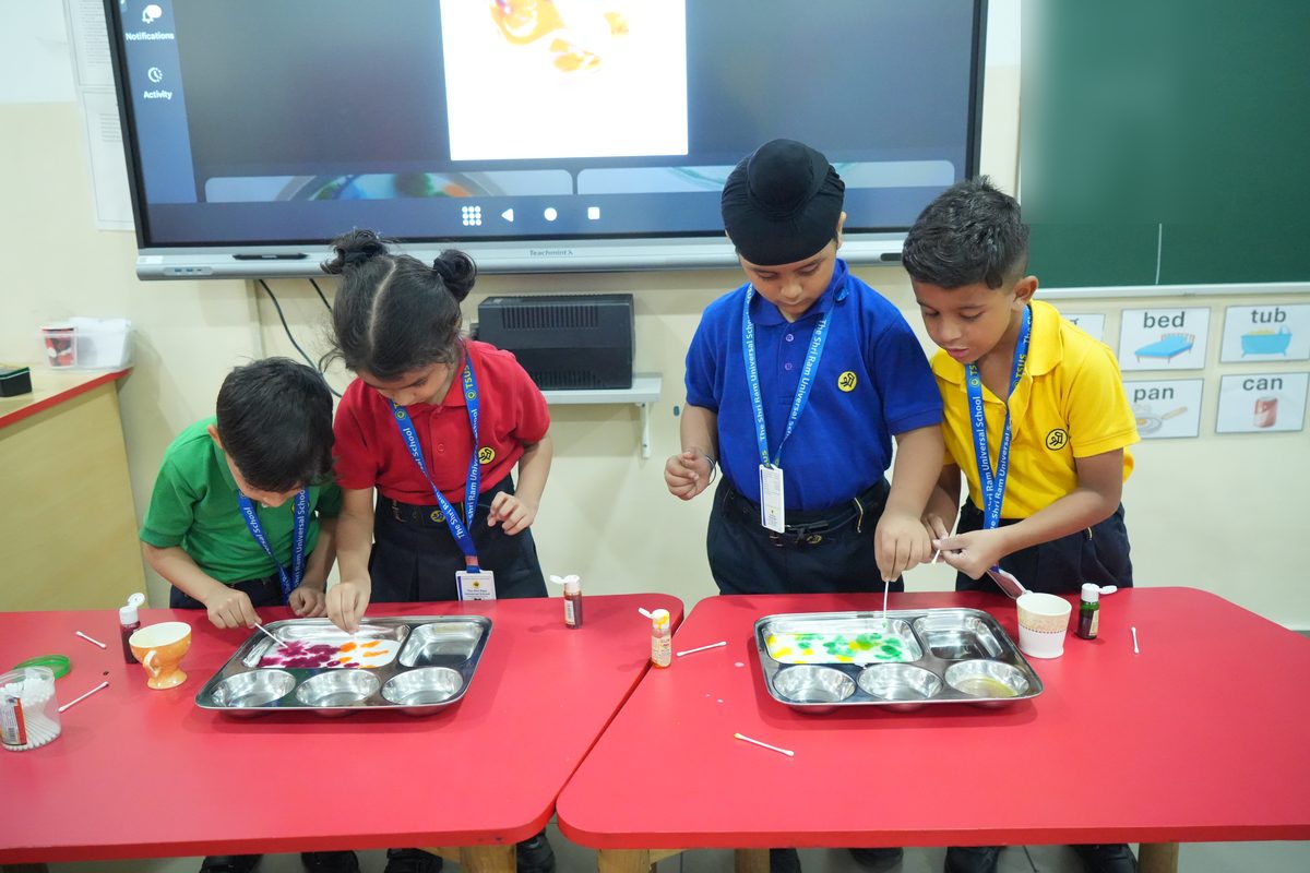 Four young children using cotton buds to mix drops of colour in steel trays during a Wonder Time activity at The Shri Ram Universal School, Sarabha Nagar