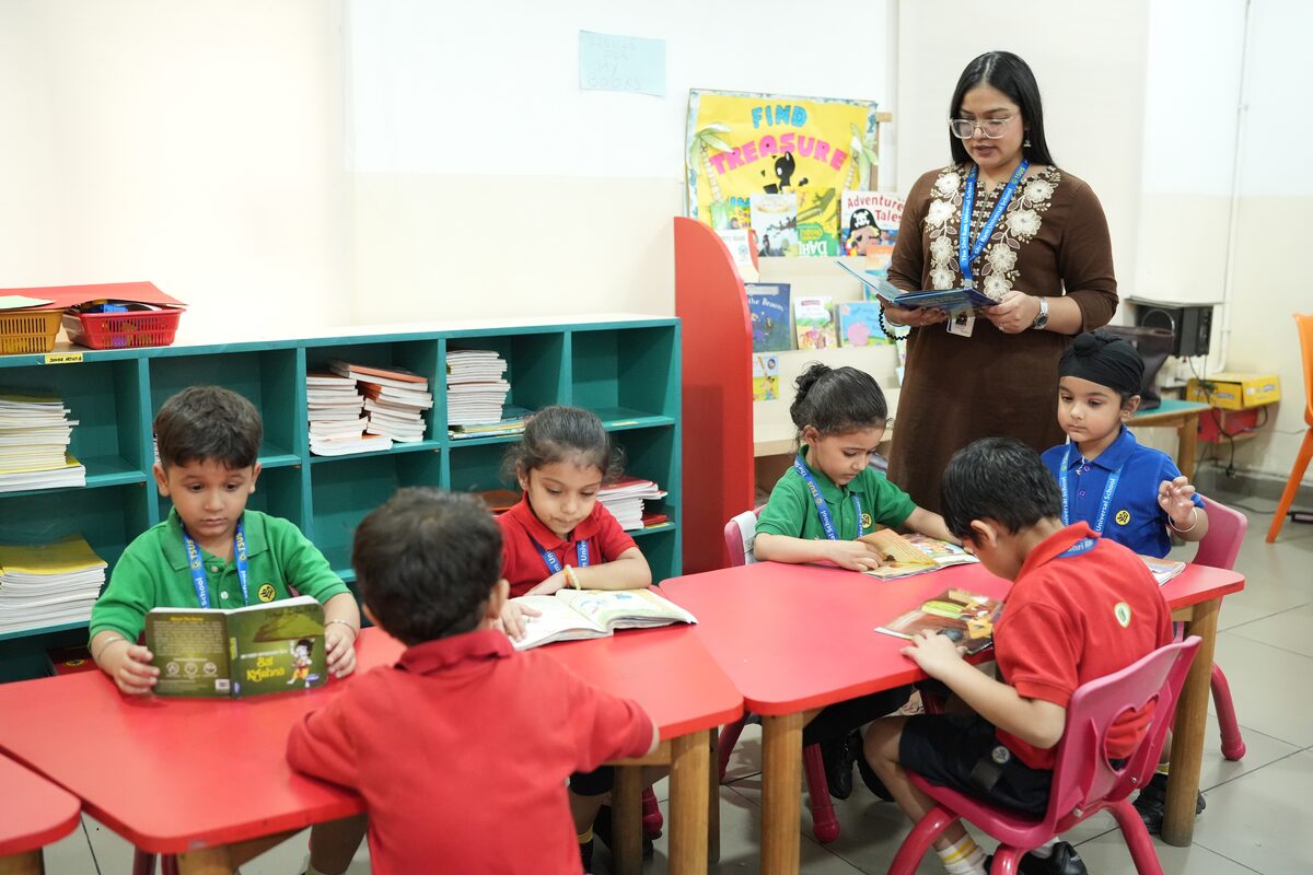 Kindergarten children reading picture books at their tables while their teacher holds up a storybook