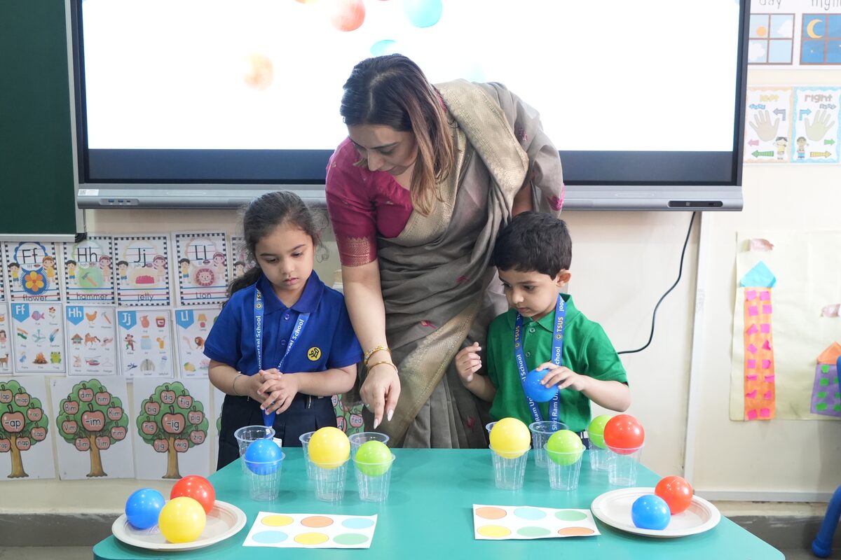 A teacher helping two Pre-Nursery children sort coloured balls into cups in their classroom