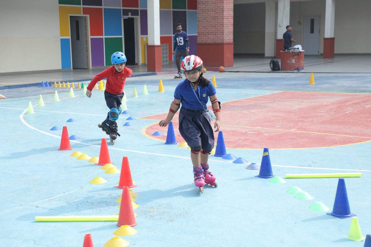 Primary children in helmets and pads skate between cones on the skating rink at TSUS Sarabha Nagar