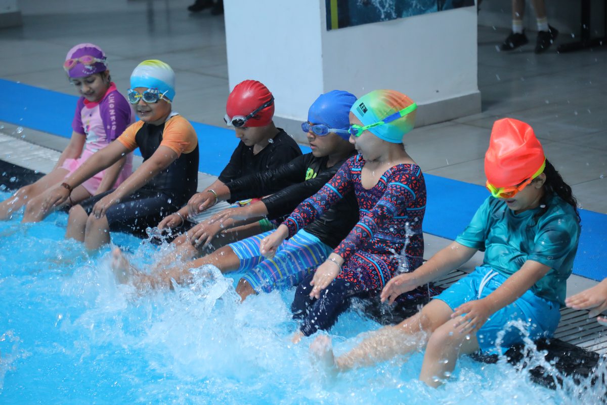 A row of students in swim caps sit on the pool edge and kick their legs in the all-weather pool at TSUS Sarabha Nagar